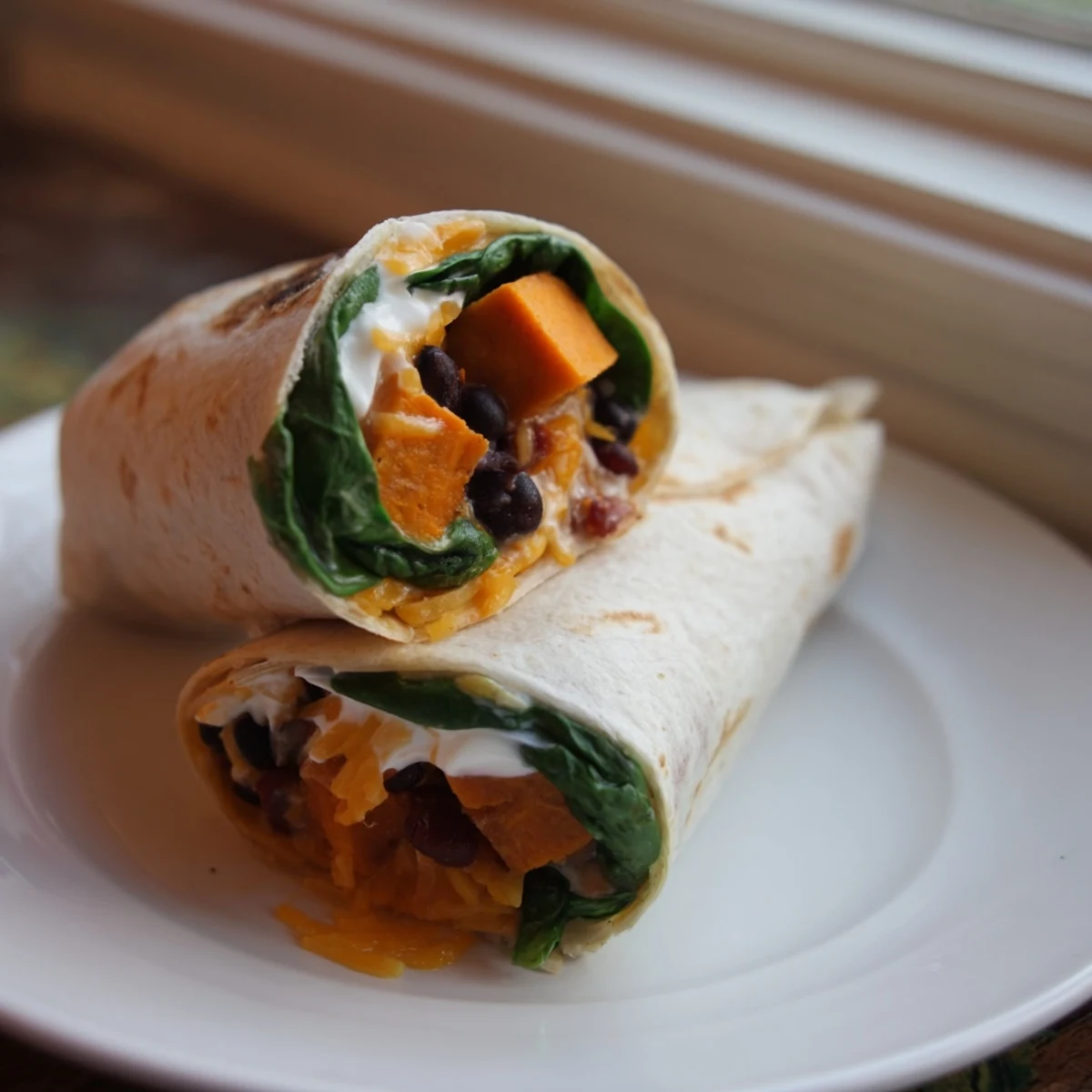 A hand holds a cut Roasted Sweet Potato and Black Bean Burrito, revealing layers of caramelized sweet potatoes, black beans, and fresh toppings with melted cheese on a rustic plate.
