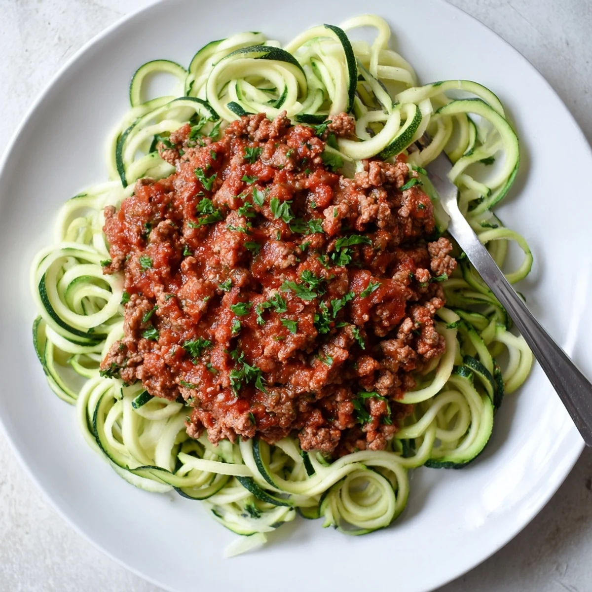 Freshly spiralized zucchini noodles topped with a simmered turkey Bolognese sauce, garnished with parsley and a pinch of red pepper flakes for a low-carb, gluten-free main dish.  