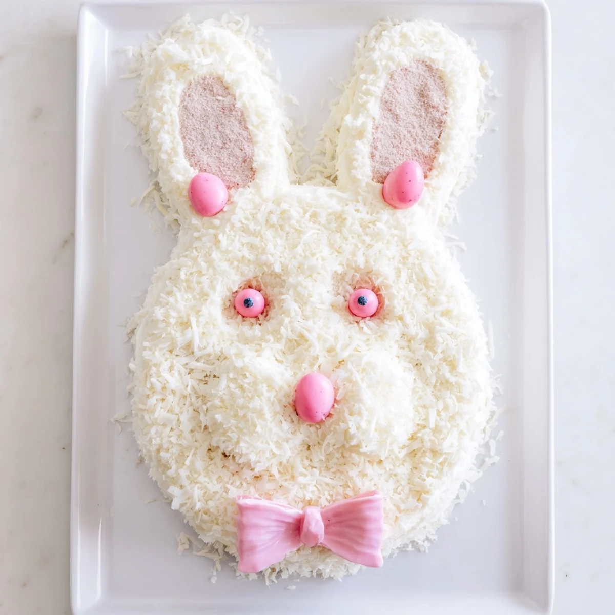 Festive spring bunny cake decorated with white coconut fur, pink tinted ears, and candy nose and eyes